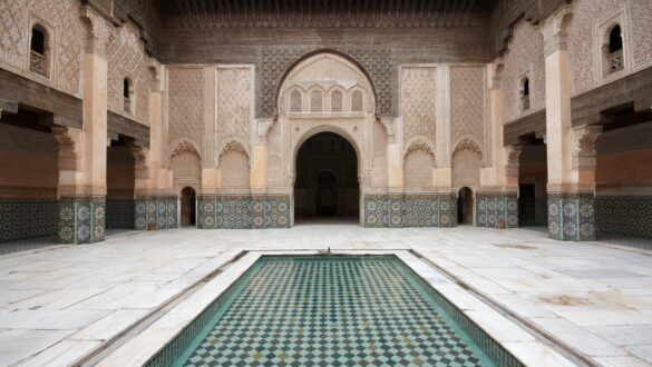 The image is a photo of the interior of Ali ben Youssef Medersa in Marrakech, Morocco, showcasing the intricate plasterwork, zellige tilework, and woodwork that are prominent features of Moroccan architecture and design influenced by Islamic art and culture.