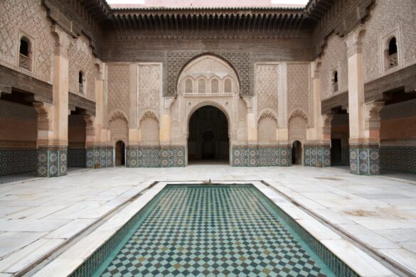 The image is a photo of the interior of Ali ben Youssef Medersa in Marrakech, Morocco, showcasing the intricate plasterwork, zellige tilework, and woodwork that are prominent features of Moroccan architecture and design influenced by Islamic art and culture.