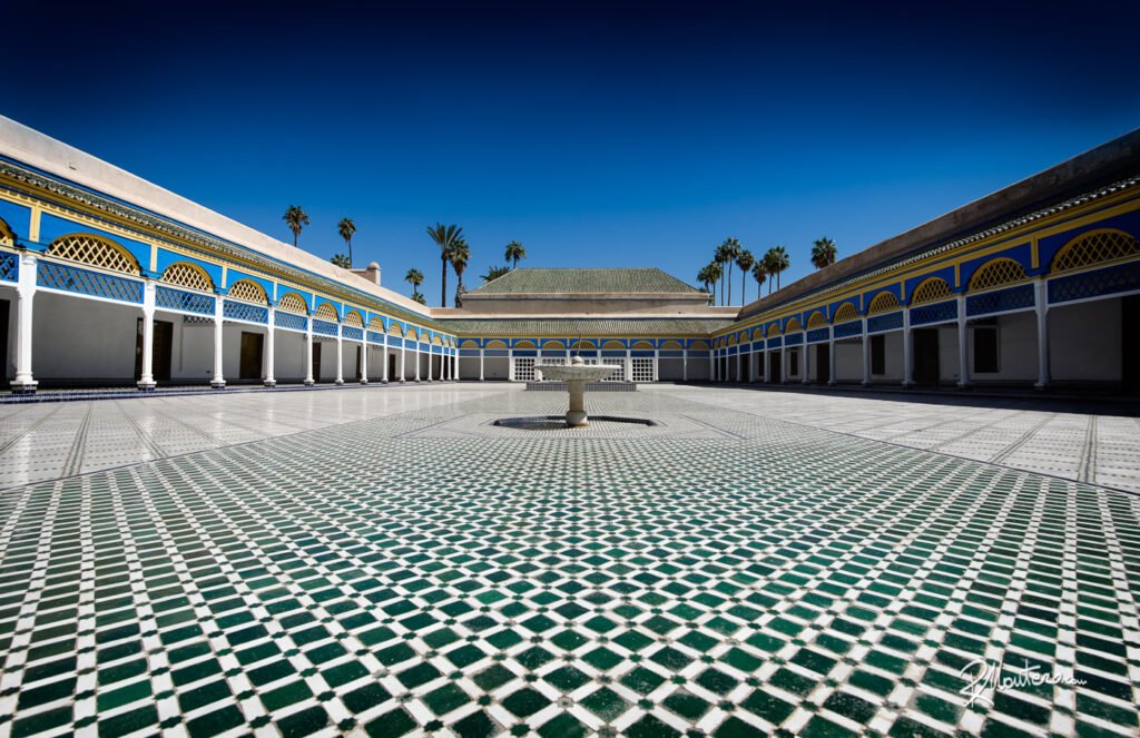 An ornate courtyard with intricate zellige tilework, stucco carvings, and lush greenery in Bahia Palace, Marrakech, showcasing the beauty of Moroccan palace architecture and design.