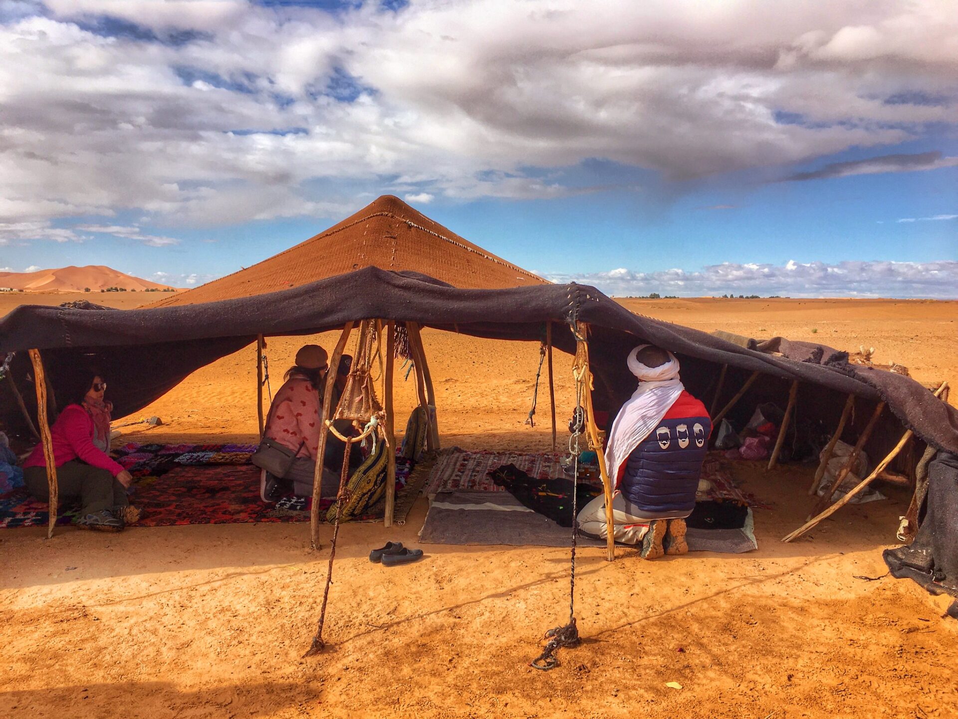 A group of people sitting on colorful carpets and cushions, drinking tea, and enjoying the view of Erg Chebbi dunes in the background at a Berber tribe camp in the Sahara Desert, Morocco.