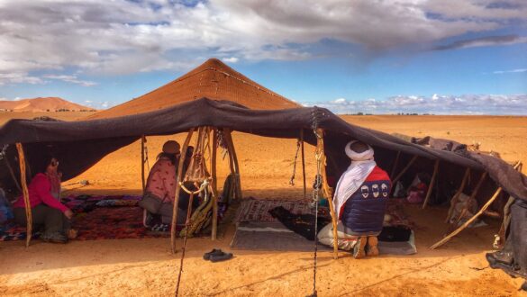 A group of people sitting on colorful carpets and cushions, drinking tea, and enjoying the view of Erg Chebbi dunes in the background at a Berber tribe camp in the Sahara Desert, Morocco.