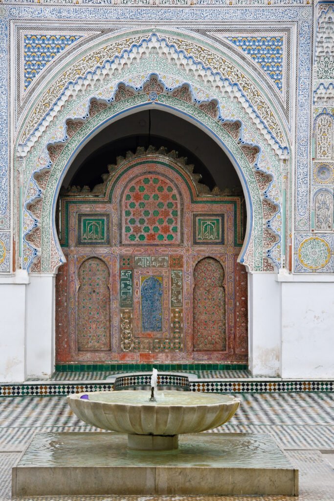 A fountain in the courtyard of the Al-Qarawiyyin Mosque and University in Fez, Morocco. The courtyard is surrounded by traditional Moroccan architecture with intricate tilework and archways. The fountain is the central focal point of the courtyard.