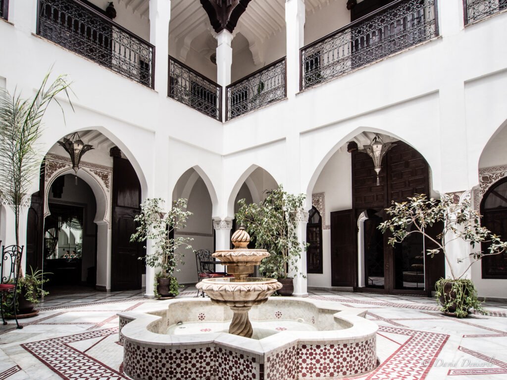 Exterior view of Riad Nasreen, a traditional Moroccan house with a central courtyard and a small pool, located in Marrakech. The image shows a wooden door leading to the courtyard, with intricate tilework and plasterwork covering the walls.Exterior view of Riad Nasreen, a traditional Moroccan house with a central courtyard and a small pool, located in Marrakech. The image shows a wooden door leading to the courtyard, with intricate tilework and plasterwork covering the walls.