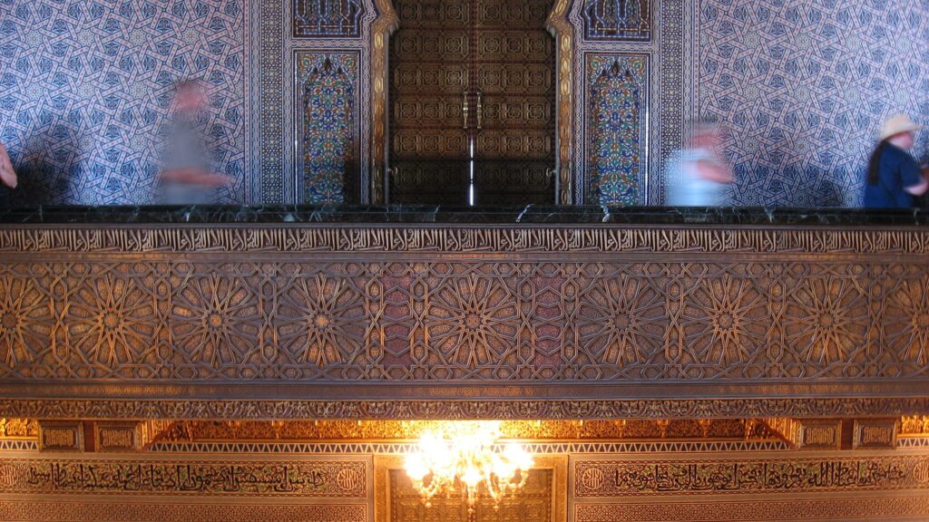 Interior of the Mausoleum of Mohammed V in Morocco, showcasing intricate traditional Moroccan architectural details including ornate tilework, carved plaster, and painted wood. The space features a high ceiling with a large chandelier, and a central area with a tomb adorned with colorful floral arrangements and candles.