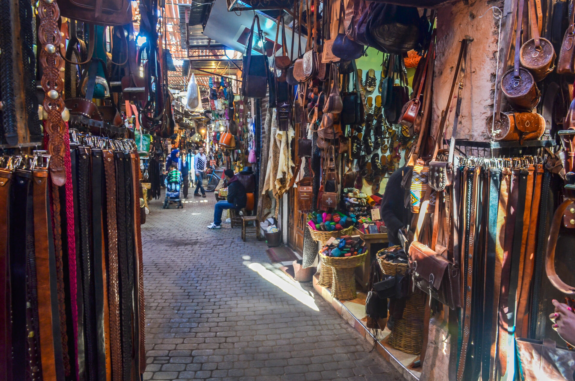 A vibrant and busy scene of the souks in Marrakech, Morocco. Stalls and shops selling various items such as textiles, ceramics, spices, and jewelry are seen in the narrow alleys, while people walk and shop around them.