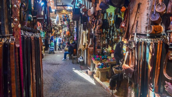 A vibrant and busy scene of the souks in Marrakech, Morocco. Stalls and shops selling various items such as textiles, ceramics, spices, and jewelry are seen in the narrow alleys, while people walk and shop around them.