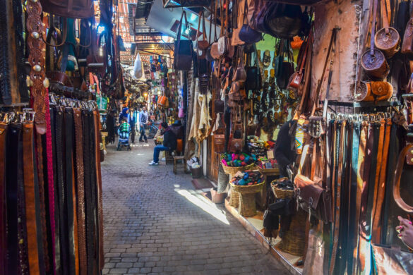 A vibrant and busy scene of the souks in Marrakech, Morocco. Stalls and shops selling various items such as textiles, ceramics, spices, and jewelry are seen in the narrow alleys, while people walk and shop around them.