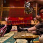 A Berber man wearing traditional clothing is serving tea to guests, representing the warmth and generosity of Moroccan hospitality. The man is standing in front of a rug with a decorative pattern, and the tea is served in a small glass on a silver tray with mint leaves on top.