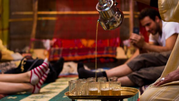 A Berber man wearing traditional clothing is serving tea to guests, representing the warmth and generosity of Moroccan hospitality. The man is standing in front of a rug with a decorative pattern, and the tea is served in a small glass on a silver tray with mint leaves on top.