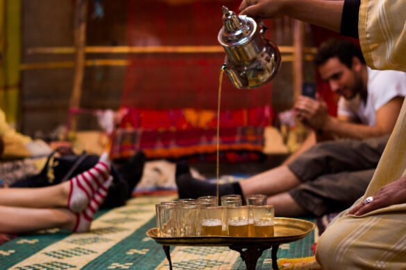 A Berber man wearing traditional clothing is serving tea to guests, representing the warmth and generosity of Moroccan hospitality. The man is standing in front of a rug with a decorative pattern, and the tea is served in a small glass on a silver tray with mint leaves on top.