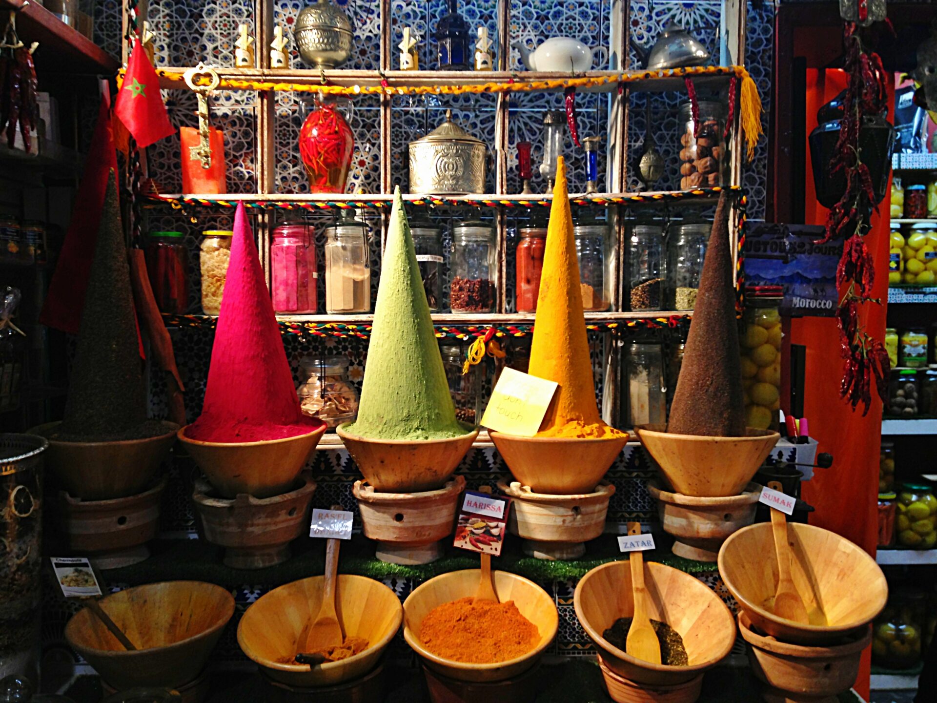 A colorful display of Moroccan spices arranged in open baskets and jars in a market stall or store. Various spices can be seen such as saffron, turmeric, paprika, and cumin. The stall is adorned with traditional Moroccan lamps and textiles, adding to the overall ambiance.