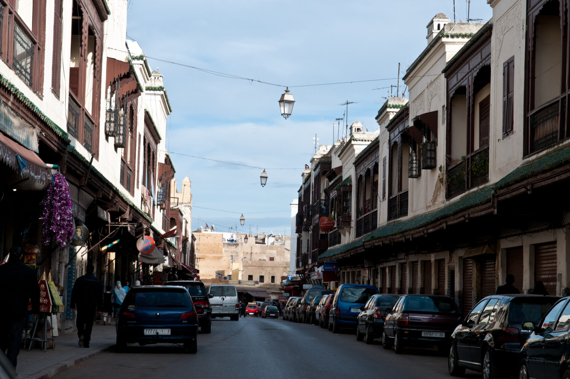  A view of the narrow alleyways and colorful buildings in the Jewish Quarter of Fes, Morocco. The photo shows people walking and exploring the historic neighborhood, with traditional Moroccan architecture visible in the background.