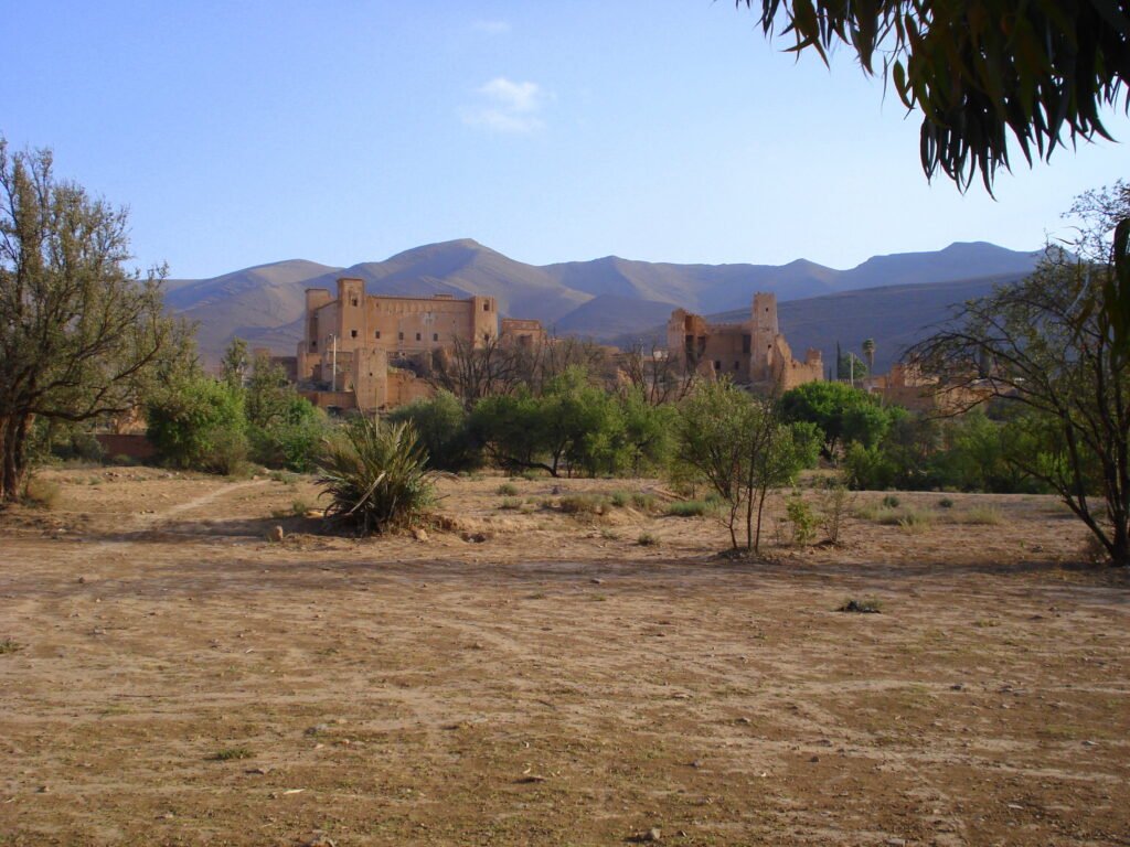 A distant view of Kasbah of Glaoui in Taliouine, Morocco, known as a gateway to the High Atlas Mountains and other treasures of Morocco. The Kasbah is a fortified citadel made of red clay and straw, with tall towers and ornate details.