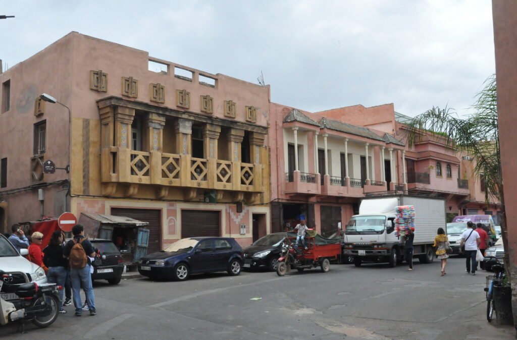 The image appears to depict a view of the Mellah neighborhood in Marrakech, with bustling streets lined with colorful buildings and storefronts. The neighborhood is filled with people going about their daily activities and various goods and products for sale, including rugs and textiles, spices, and fresh produce. The architecture and design of the buildings reflect the rich cultural heritage and history of the Mellah, which has been an important center for Jewish life in Marrakech for centuries.