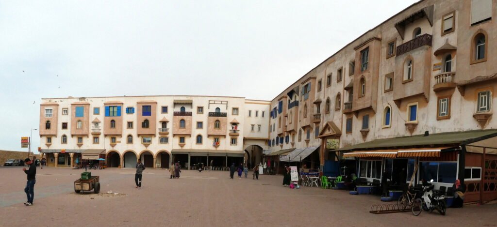 The image appears to show a street in Essaouira's Jewish quarter, with buildings on either side and a clear blue sky above. People can be seen walking along the street, and some shops and market stalls are visible.