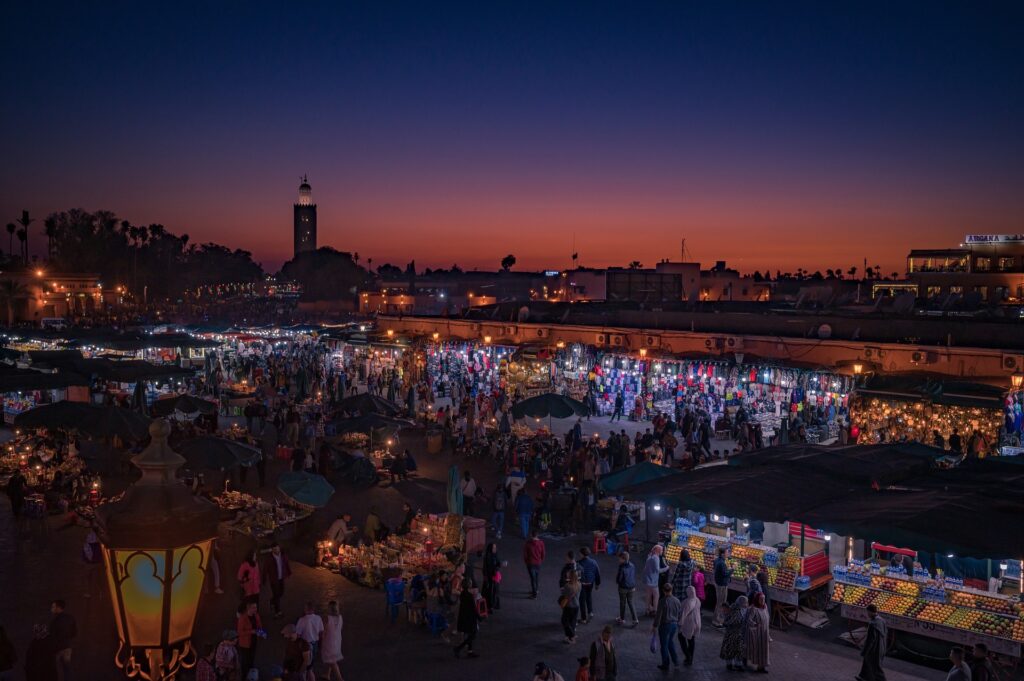 People gathered in Jemaa el-Fnaa Square at night, with food vendors, musicians, and street performers in the background.