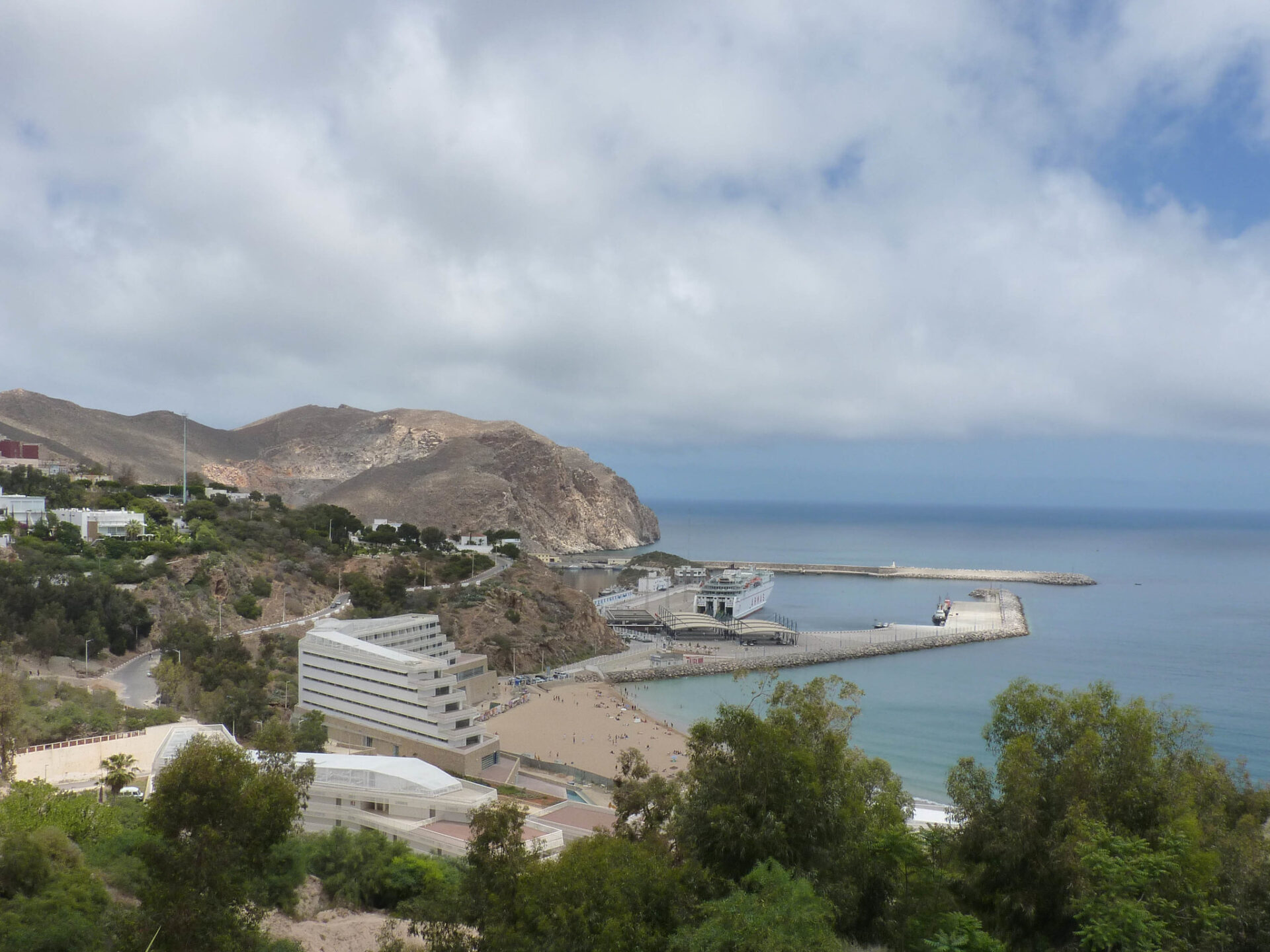 A panoramic view of Al Hoceïma, a hidden gem, featuring pristine sandy beaches, turquoise waters, and a charming coastal town. The image showcases a picturesque scene with a row of colorful buildings lining the shoreline, complemented by palm trees and a clear blue sky, inviting visitors to relax and indulge in the beauty of this coastal paradise.