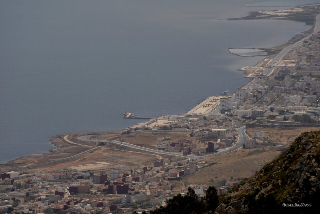 A view of the Nador waterfront, known as Paseo marítimo de Nador, with a newly constructed road extending from Mount Ubayo. The image captures the expansive promenade along the coast, lined with palm trees, streetlights, and modern buildings. The road stretches into the distance, leading towards the majestic Mount Ubayo, creating a picturesque blend of urban and natural elements in Nador.