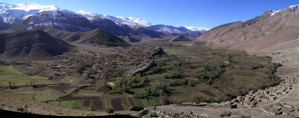 A breathtaking panoramic view of the snow-covered Aït Bouguemez Valley in the High Atlas Mountains. The image captures the picturesque landscape with terraced hills and white snow-capped mountains, showcasing the natural beauty of this hidden gem in Morocco