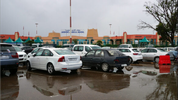 A rainy day at Agadir Airport with the taxi station visible in the foreground and the airport entry hall in the background, showcasing wet pavement and a cloudy sky.