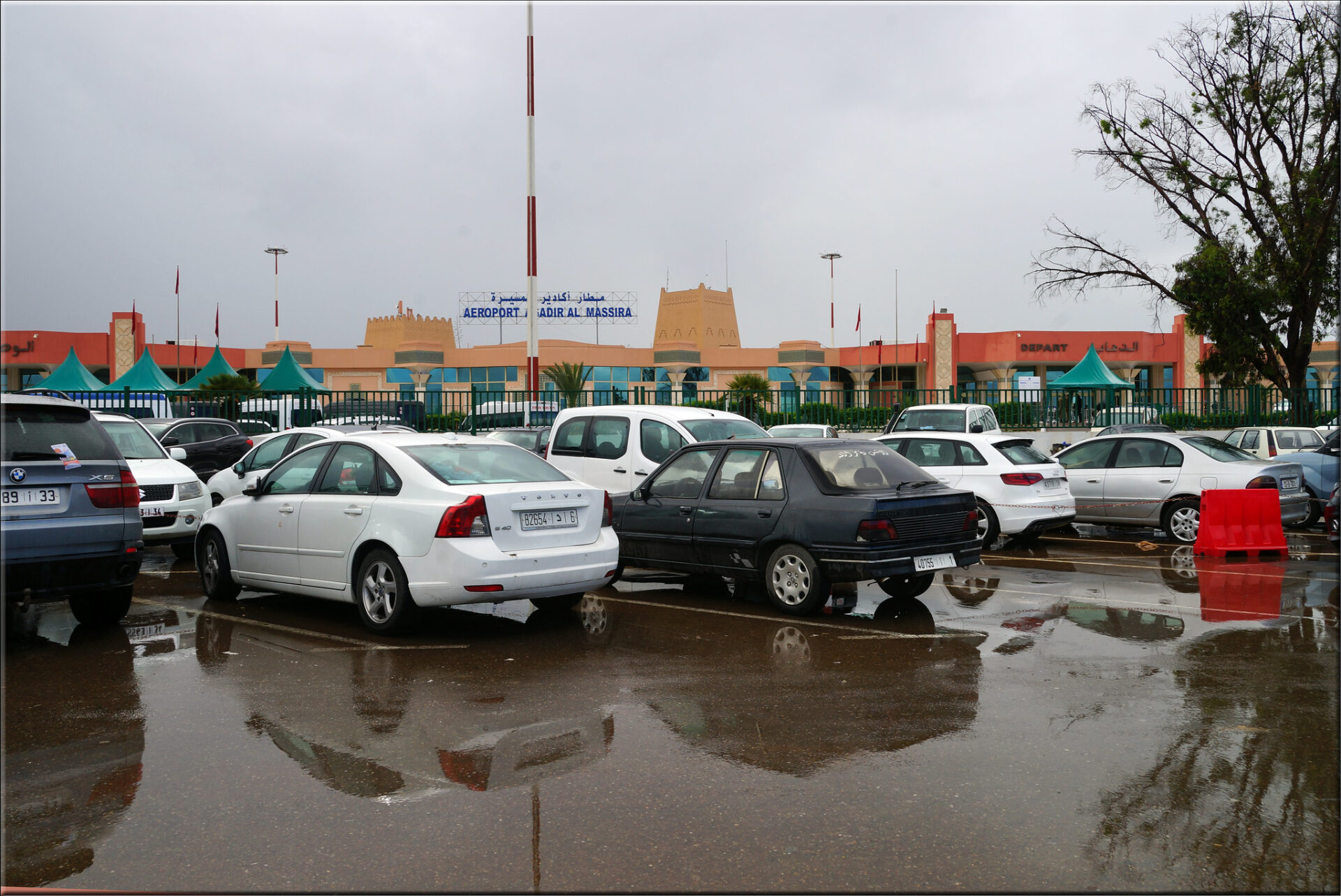 A rainy day at Agadir Airport with the taxi station visible in the foreground and the airport entry hall in the background, showcasing wet pavement and a cloudy sky.